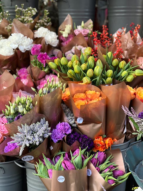 A vibrant display of freshly arranged flowers including white and pink carnations, purple statice, green tulip buds, and orange rose petals, all wrapped in brown craft paper. The bouquets are grouped 