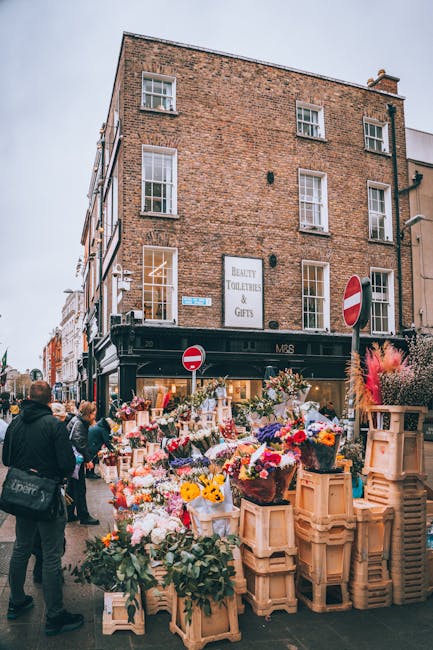 The image depicts an outdoor flower market on a bustling street corner, with numerous bouquets and floral arrangements displayed on wooden crates and shelves. The flowers include vibrant roses, tulips