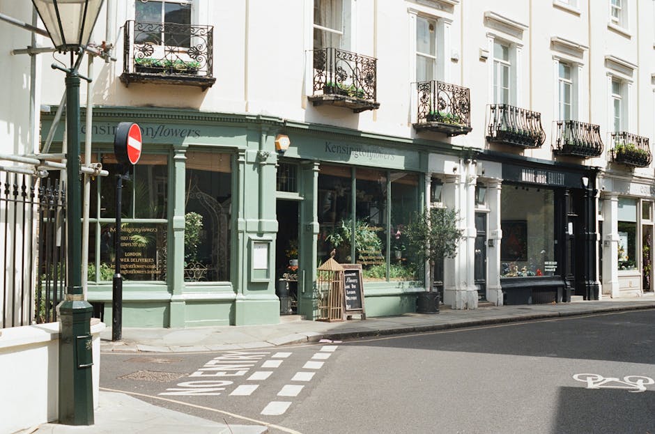 A charming flower shop situated on Marylebone High Street, featuring a pastel green façade with large glass windows displaying vibrant floral arrangements inside. The shop's name is partially visible