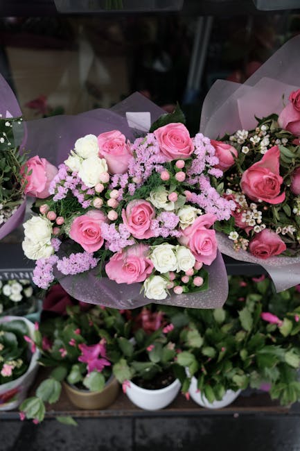 A floral arrangement featuring a bouquet of fresh pink roses, white carnations, and light purple astilbe, all wrapped in delicate lavender tissue paper with a subtle shimmer. The flowers are arranged