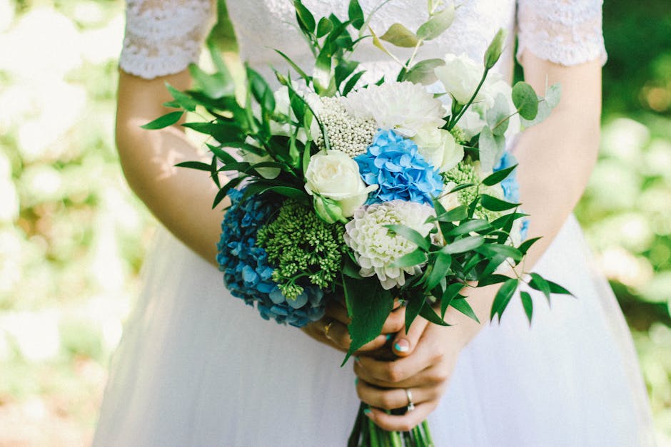 A person wearing a white lace dress holding a floral bouquet with white roses, blue hydrangeas, white carnations, and various green foliage. The arrangement appears fresh with vibrant colors and a tex