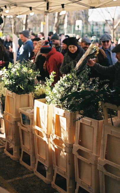 A group of people browsing floral arrangements at an outdoor flower market, with several large wooden crates filled with fresh green foliage and flowering plants. The flowers vary in shades of green,
