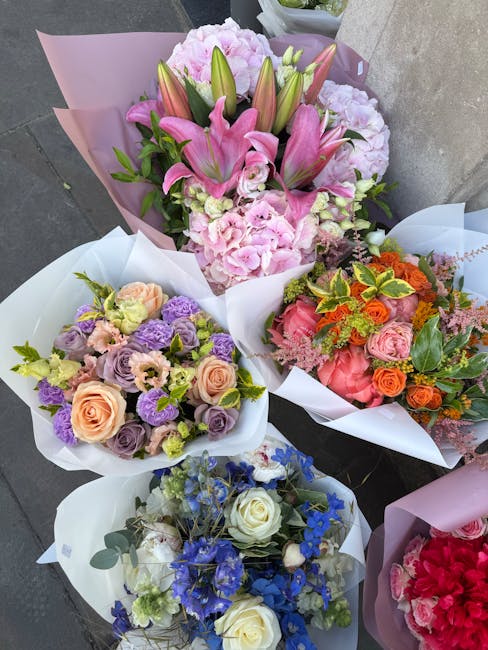 Four beautifully arranged floral bouquets placed outdoors against a neutral background. The largest bouquet features pink lilies, pale pink peonies, and green-tipped leaves wrapped in light purple tis
