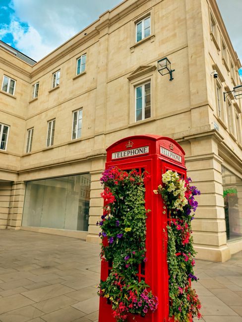 A traditional red British telephone booth adorned with an abundance of colorful flowers, including pink, purple, yellow, and white blooms, arranged in hanging baskets and climbing clusters along its s