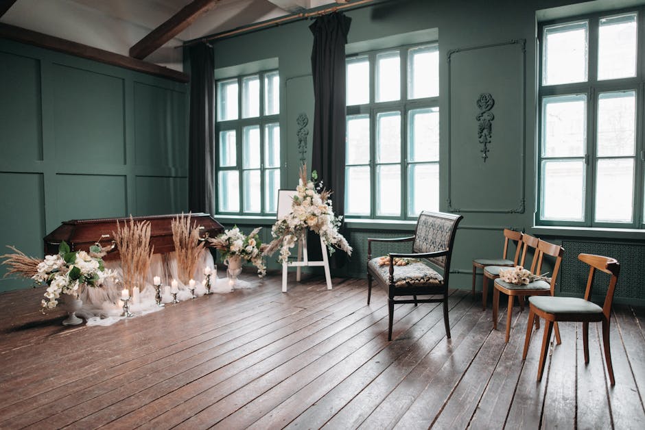 A floral arrangement featuring white roses, peonies, and orchids with accents of dried pampas grass and greenery, displayed on a white easel in a softly lit room with large multi-pane windows. Two sma