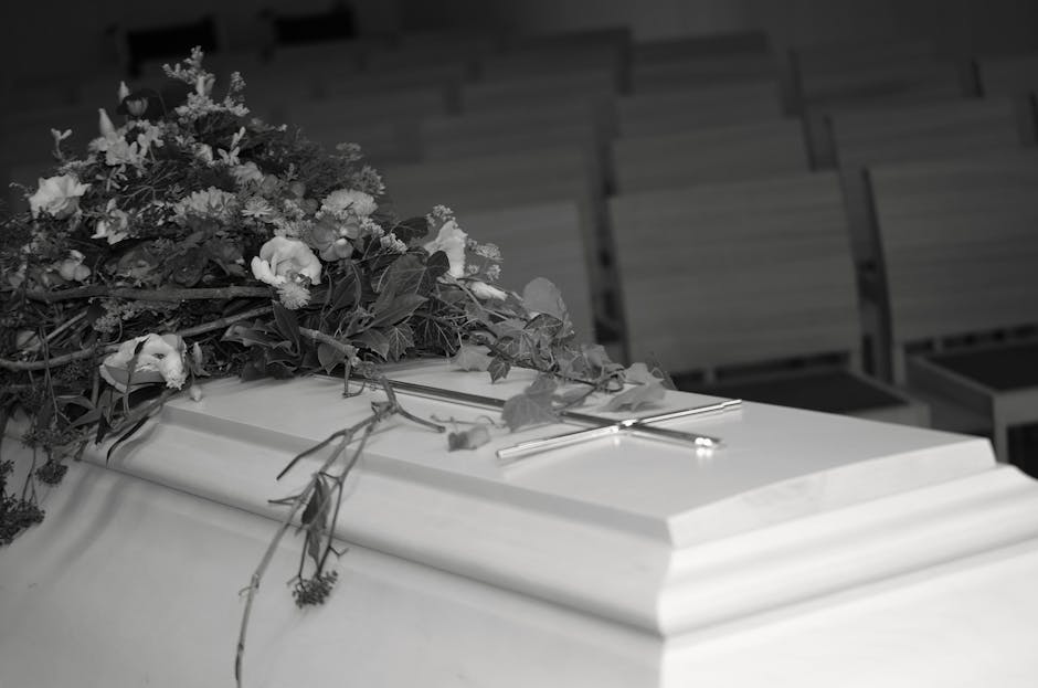 A floral arrangement featuring a mixture of fresh white roses, lilies, and greenery, intricately arranged on top of a closed white casket with floral leaves and stems cascading over the sides. The bou
